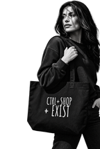 A woman in a black sweatshirt and jeans carries a black canvas tote bag with the white text 'CTRL+SHOP + EXIST' 